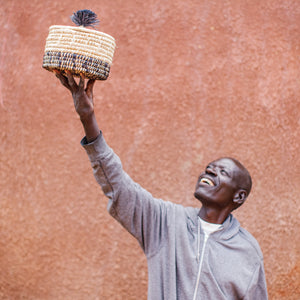 JustOne's natural basket with black details on bottom and lid with a black pompom handle, handcrafted in Uganda