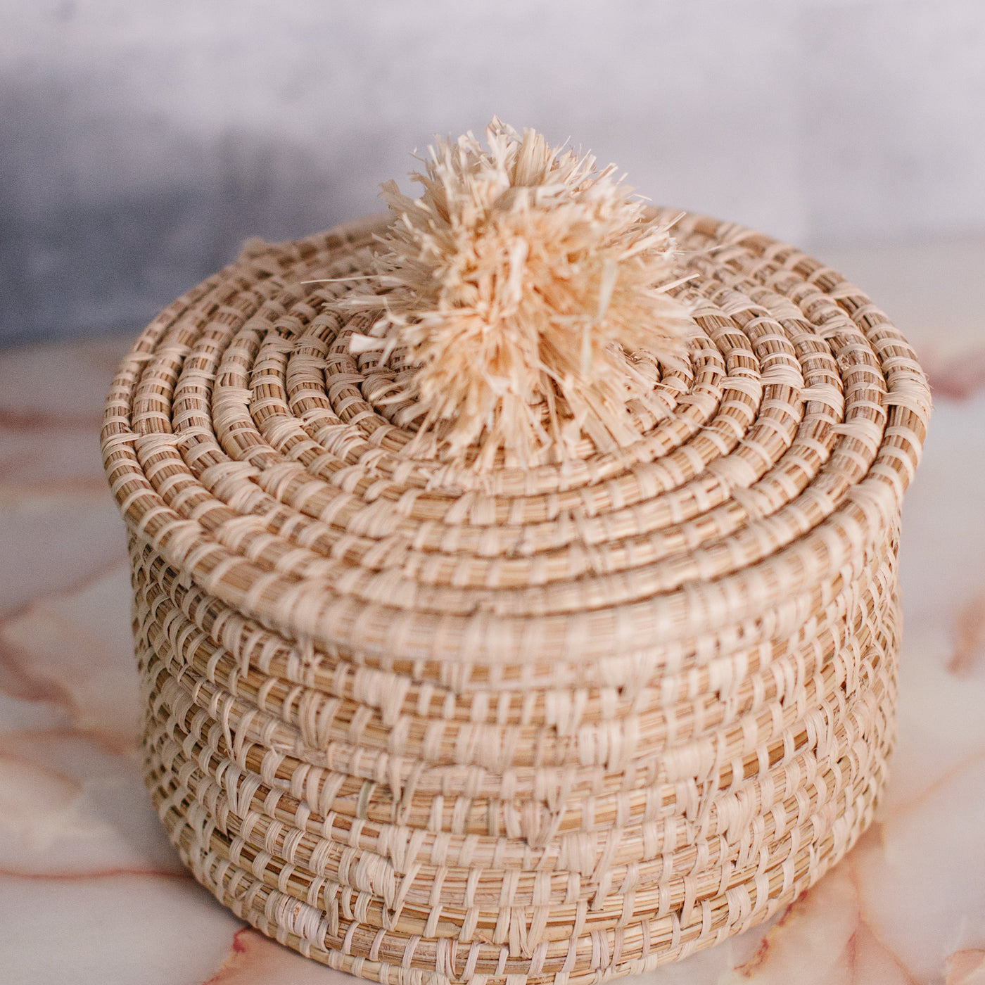 JustOne's tan basket and lid with a tan pompom handle, handcrafted in Uganda