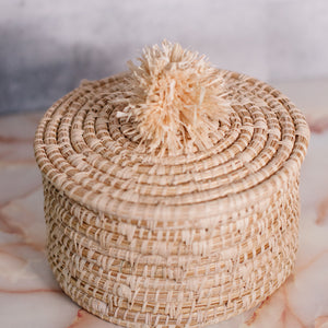 JustOne's tan basket and lid with a tan pompom handle, handcrafted in Uganda