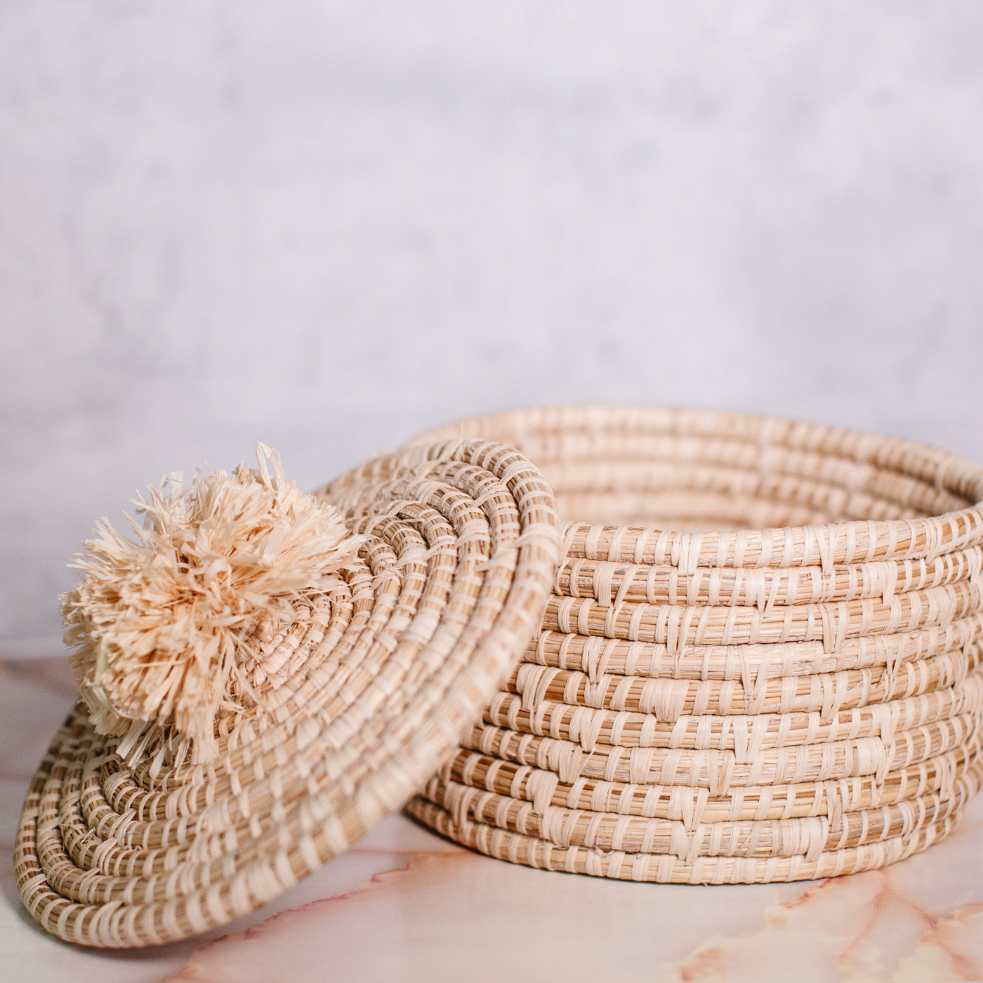 JustOne's tan basket and lid with a tan pompom handle, handcrafted in Uganda